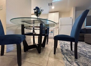 Un comedor con mesa de vidrio, sillas oscuras, un jarrón con flores y una cocina al fondo.