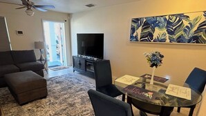 Ein Wohnzimmer mit einer Couch, Hocker, Couchtisch und Stühlen. Es gibt einen Flachbildfernseher, einen Deckenventilator und ein Bild an der Wand.