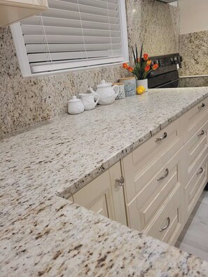 Una cocina con encimera de granito, armarios blancos, ventana con persianas y una pequeña maceta con flores naranjas.