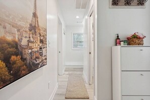 Un pasillo con un mural grande de un paisaje urbano, un armario blanco con artículos de aseo, y una ventana con persianas.