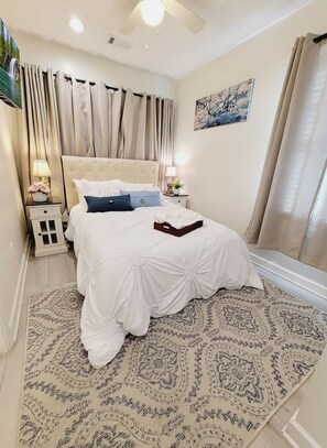 Un dormitorio con cama, mesitas de noche, ventilador de techo, ventana con cortinas y una obra de arte colgada en la pared.