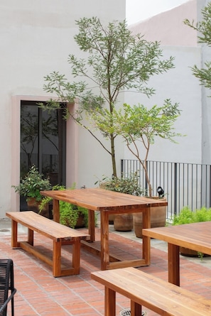 Une terrasse extérieure avec des tables et des bancs en bois, entourée de plantes en pot et d’un arbre.