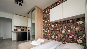 A bedroom with a bed, a wooden wardrobe, a kitchen area, and a floral wallpaper. - Praga-Południe Chic Studio (Warszawa)