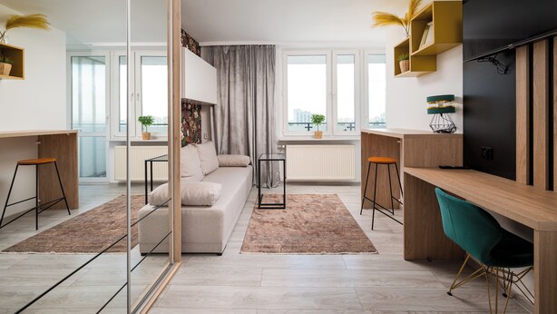 A modern living room with a sofa, a coffee table, a desk, and a TV. There are windows with curtains, a radiator, and a plant on the windowsill. - Praga-Południe Chic Studio (Warszawa)