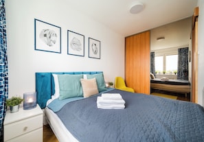 Een slaapkamer met een bed, een nachtkastje, een lamp, een raam en een badkamer zichtbaar door een geopende deur.
