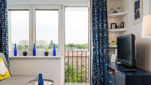 A hotel room with a bed, a TV, a small table, a window with curtains, and a balcony with potted plants. - Warszawa Wola Cozy Apartment (Warszawa)