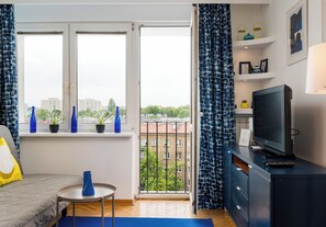 Ein Hotelzimmer mit Bett, Fernseher, kleinem Tisch, Fenster mit Vorhängen und Balkon mit Blumentöpfen.