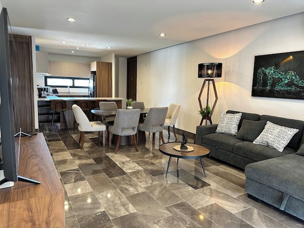 Un salon moderne avec un canapé gris, des fauteuils assortis, une table basse et un coin repas, la cuisine étant visible au fond.