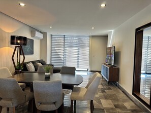 A modern living room with a dining area, a flat-screen TV, and a large window with blinds. - Stylish 2BR Apartment at Torre Bermeo (Zapopan)