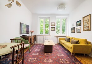 Un salon avec une table en bois, un fauteuil vert, un canapé jaune, un tapis à motifs et de grandes fenêtres.
