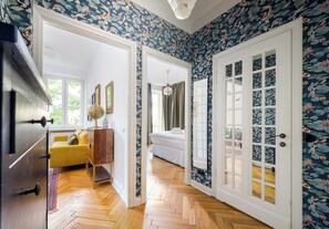 Une pièce avec un parquet en chevrons, un canapé jaune, une armoire en bois et un lit doté d’une tête de lit blanche.