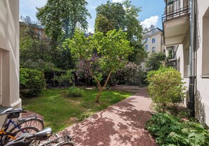 Un chemin pavé dans une cour, avec des vélos garés sur le côté gauche, le tout entouré de verdure et de bâtiments.