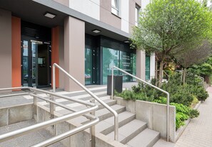 Entrada d'un edifici amb escala, baranes metàl·liques i una petita zona de jardí amb plantes.