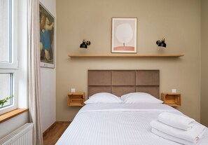 Een netjes opgemaakt bed met witte lakens, een hoofdbord, twee nachtkastjes met lampen, een ingelijste foto aan de muur, en een raam met gordijn.