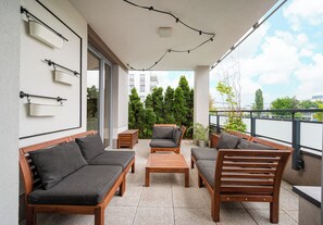 Een balkon met houten meubels, waaronder banken en een tafel, en uitzicht op bomen en gebouwen op de achtergrond.