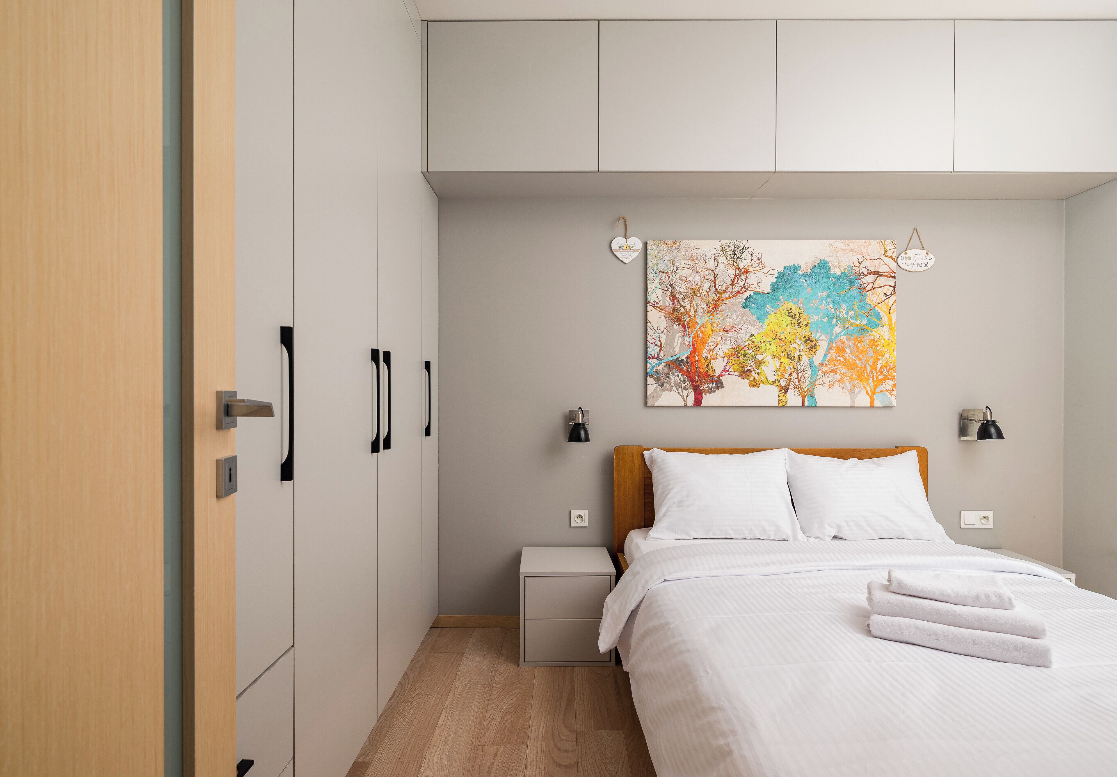 Um quarto com cama, mesas de cabeceira, um criado-mudo, uma luminária na parede e um quadro.