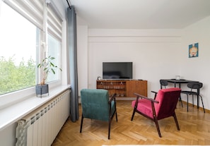 Ein Wohnzimmer mit Heizkörper, Fernseher, Holzschrank, zwei Stühlen, einem kleinen Tisch und einer Topfpflanze.