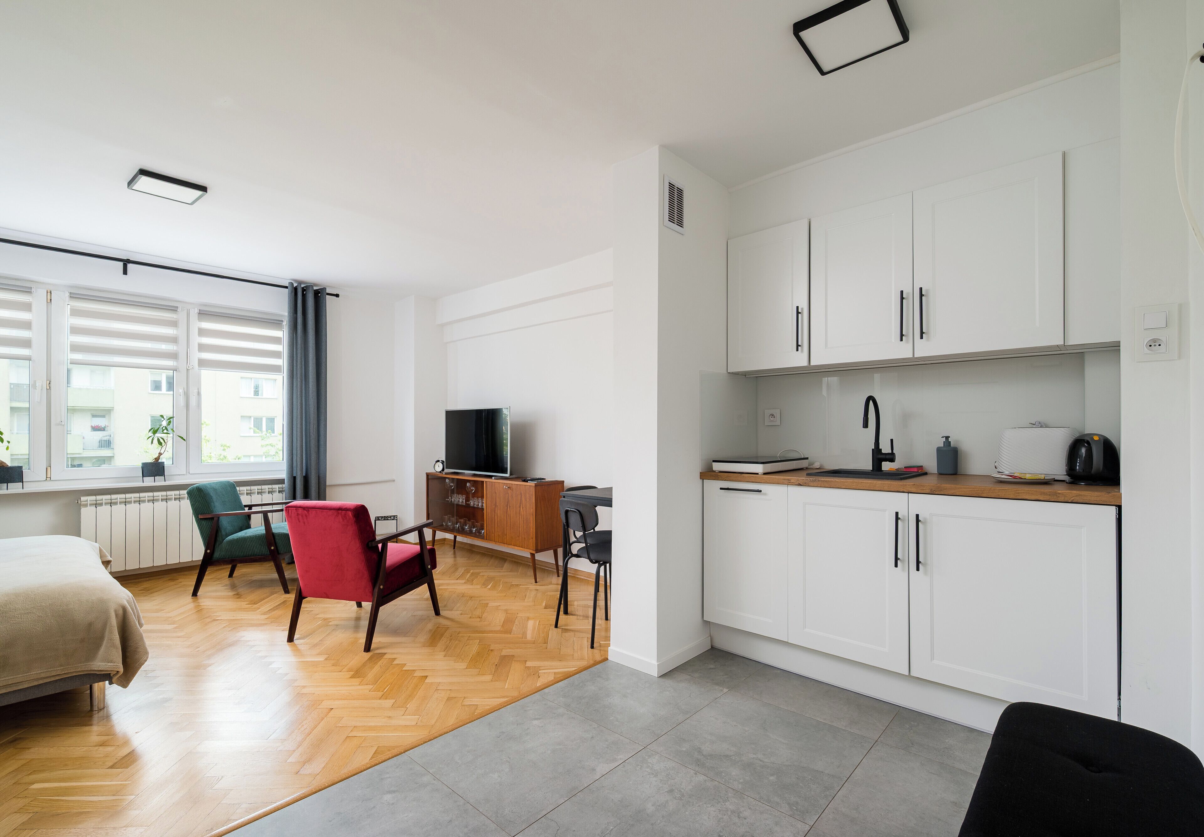 Habitació moderna amb llit, escriptori, cadira, televisió i petita cuina.