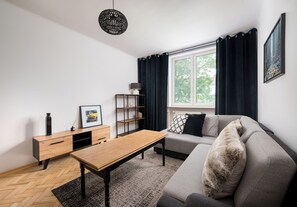 A modern living room with a sofa, a wooden coffee table, a sideboard, and a large window with curtains. - Metro Racławicka Exclusive Apartment (Warszawa)
