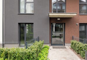 Een modern gebouw met een grijze gevel, een zwarte deur met het nummer '1A' erboven, en een klein entreegebied met een metalen hekwerk en groen.