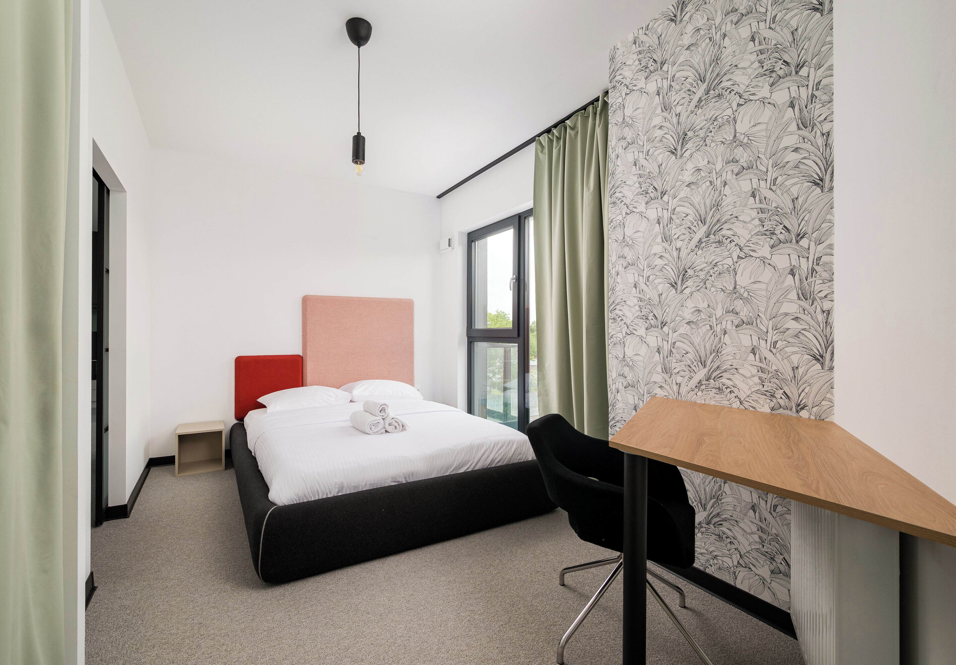 A hotel room with a bed, a desk, a chair, a window with curtains, and a wall with a floral pattern.