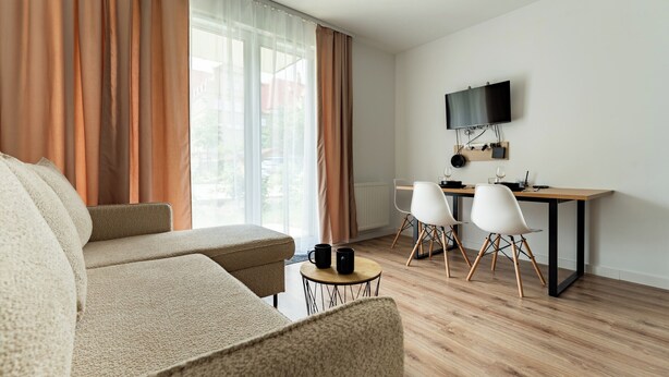 Ein modernes Wohnzimmer mit einer Couch, einem kleinen runden Tisch und einem Schreibtisch mit Stühlen. Ein Flachbildfernseher ist an der Wand montiert.