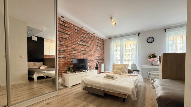 Une chambre moderne avec un mur d’accent en briques, un téléviseur à écran plat, un lit avec une tête de lit blanche, un bureau avec une chaise et une grande fenêtre avec des rideaux.