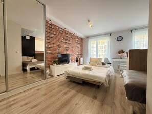 A modern bedroom with a brick accent wall, a flat-screen TV, a bed with a white headboard, a desk with a chair, and a large window with curtains. - Zakładowa Modern Living Studio (Wrocław)