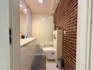 Ein modernes Badezimmer mit Waschbecken, Spiegel und Toilette in Weiß, einem kleinen Mülleimer und einem gefliesten Boden.