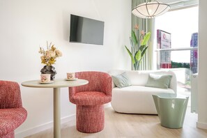 En moderne stue med en hvid sofa, røde mønstrede lænestole, et lille rundt bord med en vase og teskeer, en grøn lænestol og et TV ophængt på væggen.