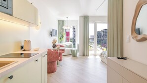 Une chambre d’hôtel moderne dotée d’un coin cuisine, d’un espace salon avec un canapé et des fauteuils, d’une grande fenêtre offrant une vue sur la ville et d’un miroir mural.