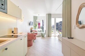 Habitación de hotel moderna con cocina, zona de estar con sofá y sillones, un amplio ventanal con vistas a la ciudad y un espejo en la pared.
