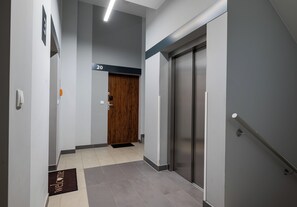 Een moderne hal met een houten deur waarop '20' staat, een deurmat en een metalen lift.