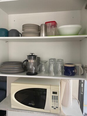 Une cuisine équipée d’un four à micro-ondes, d’une bouilloire, ainsi que de différents plats et tasses rangés sur des étagères.