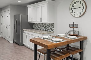 Une cuisine moderne avec des meubles blancs, un réfrigérateur en acier inoxydable, une table à manger en bois avec des bancs et une horloge murale.