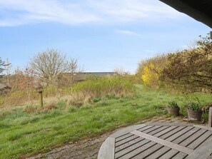 A patio area with a wooden table, potted plants, and a view of a grassy field with trees and a building in the background.