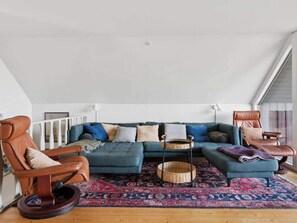 Ett vardagsrum i loftstil med en hörnsoffa, läderfåtöljer, en mönstrad matta och ett stort fönster.