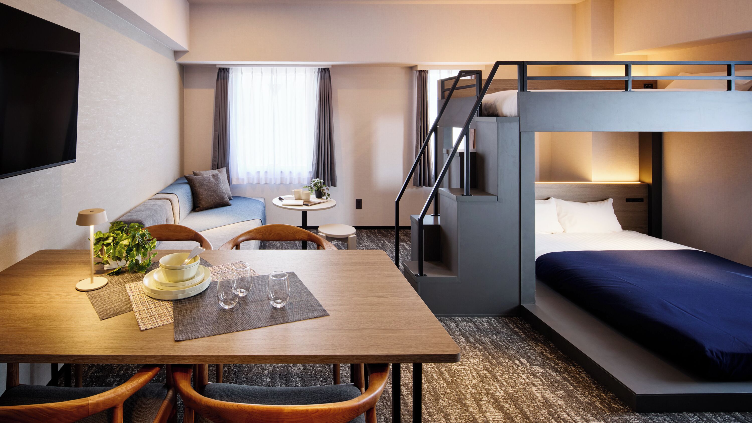 Een hotelkamer met een stapelbed, een slaapbank, een eettafel met stoelen, een bijzettafeltje met een plant en een televisie.