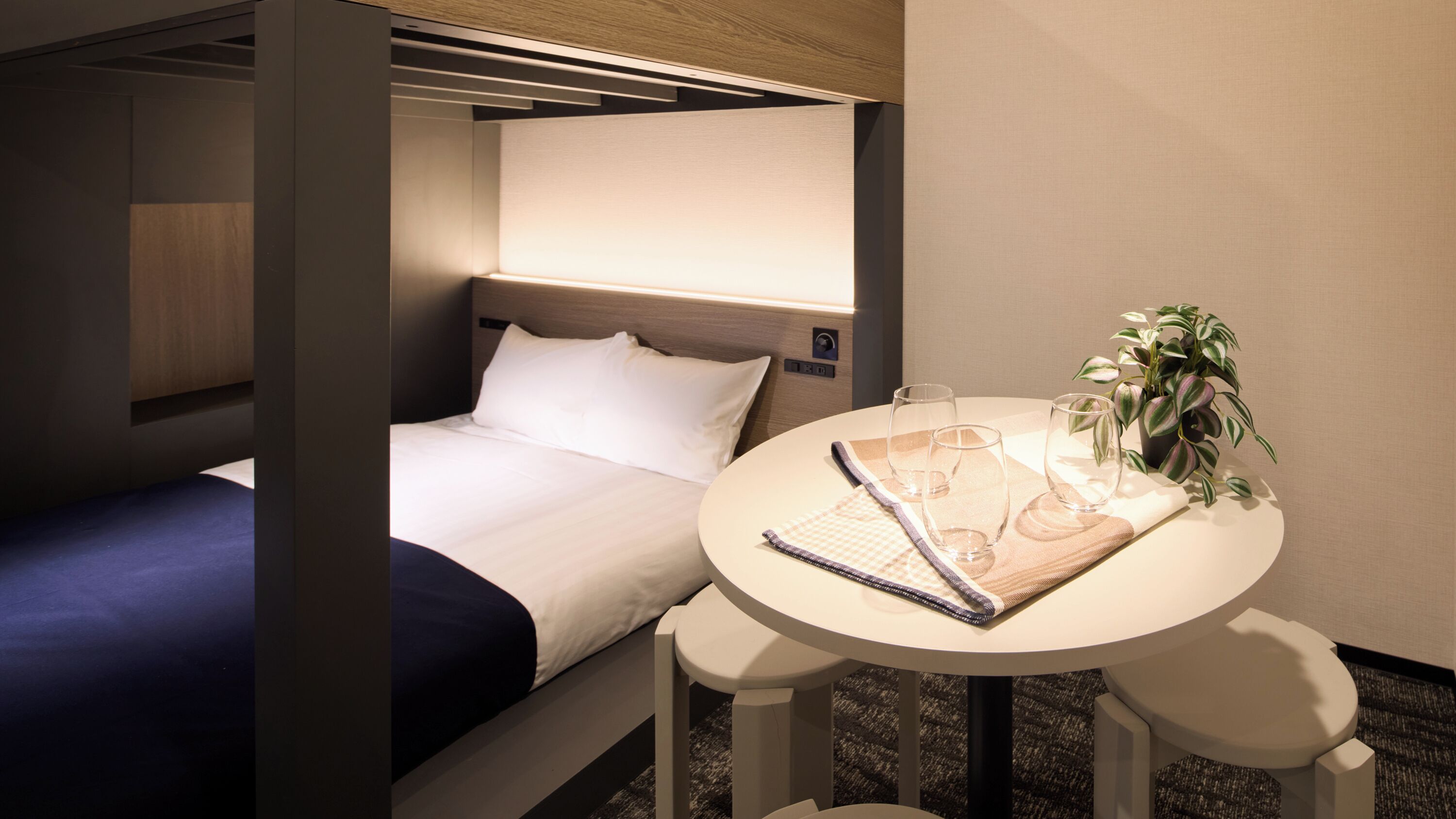 Een compacte hotelkamer met een enkel bed, een kleine tafel met glazen en een plant, en een hoofdeinde met bedieningspaneel.