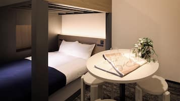 Een compacte hotelkamer met een enkel bed, een kleine tafel met glazen en een plant, en een hoofdeinde met bedieningspaneel.