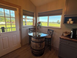 Eine Küche mit einem Holzfass als Tisch, zwei Stühlen, einer Kaffeemaschine und Blick auf ein grünes Feld durch die Fenster.