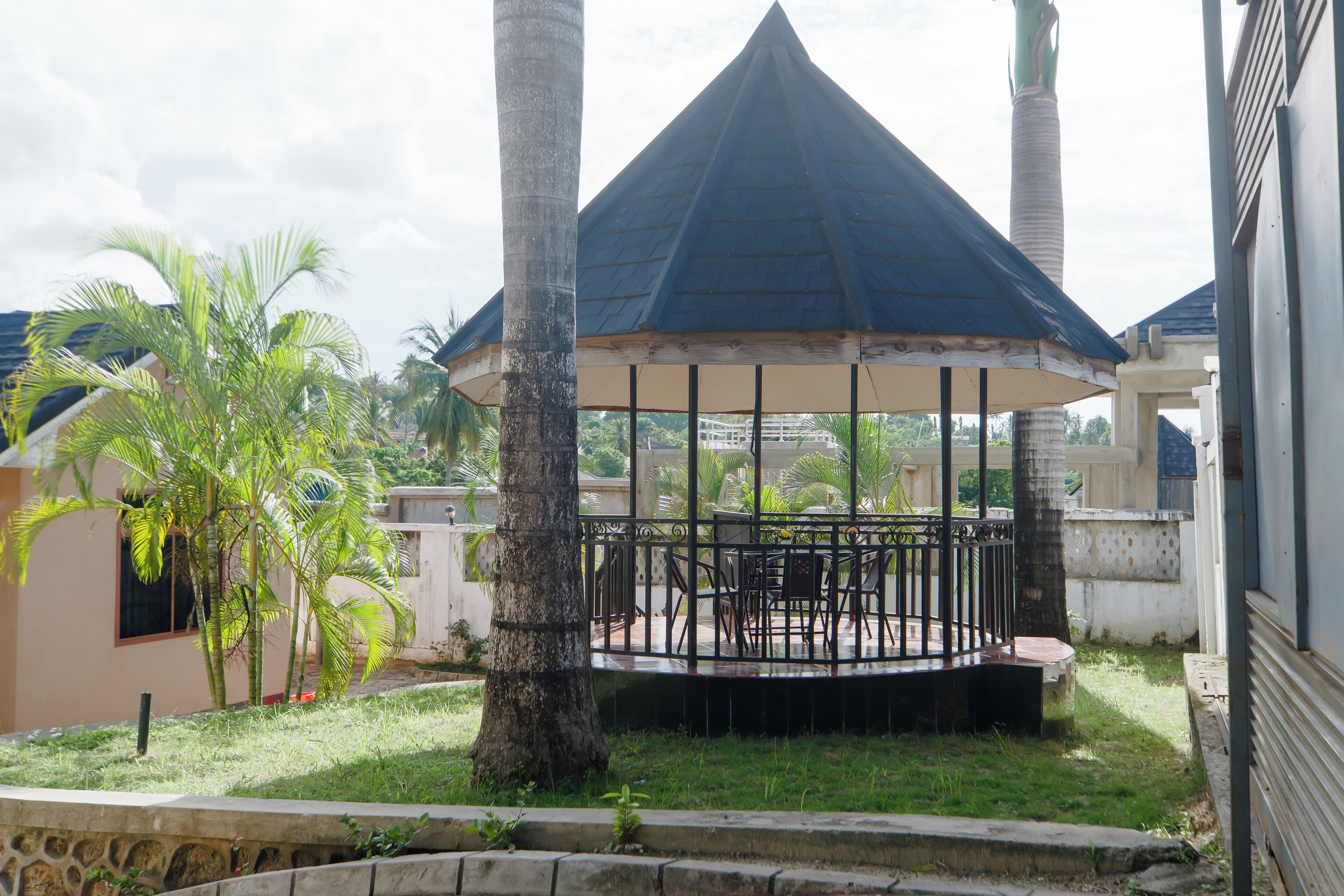 A gazebo with a blue roof and a circular table surrounded by chairs, set in a grassy area with palm trees and a building in the background.