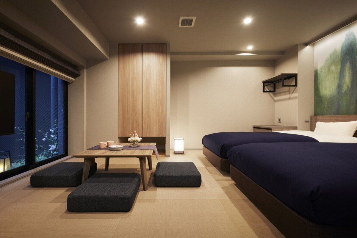 Ein modernes Hotelzimmer mit einem großen Bett, einem Schreibtisch mit Stuhl, einem kleinen Tisch mit einer Vase und einem an der Wand montierten Fernseher.