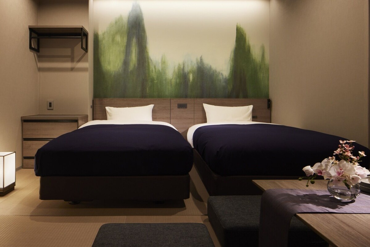 Ein Hotelzimmer mit zwei Betten, einem Nachttisch, einer Lampe, einem Bild und einem kleinen Tisch mit Blumen.