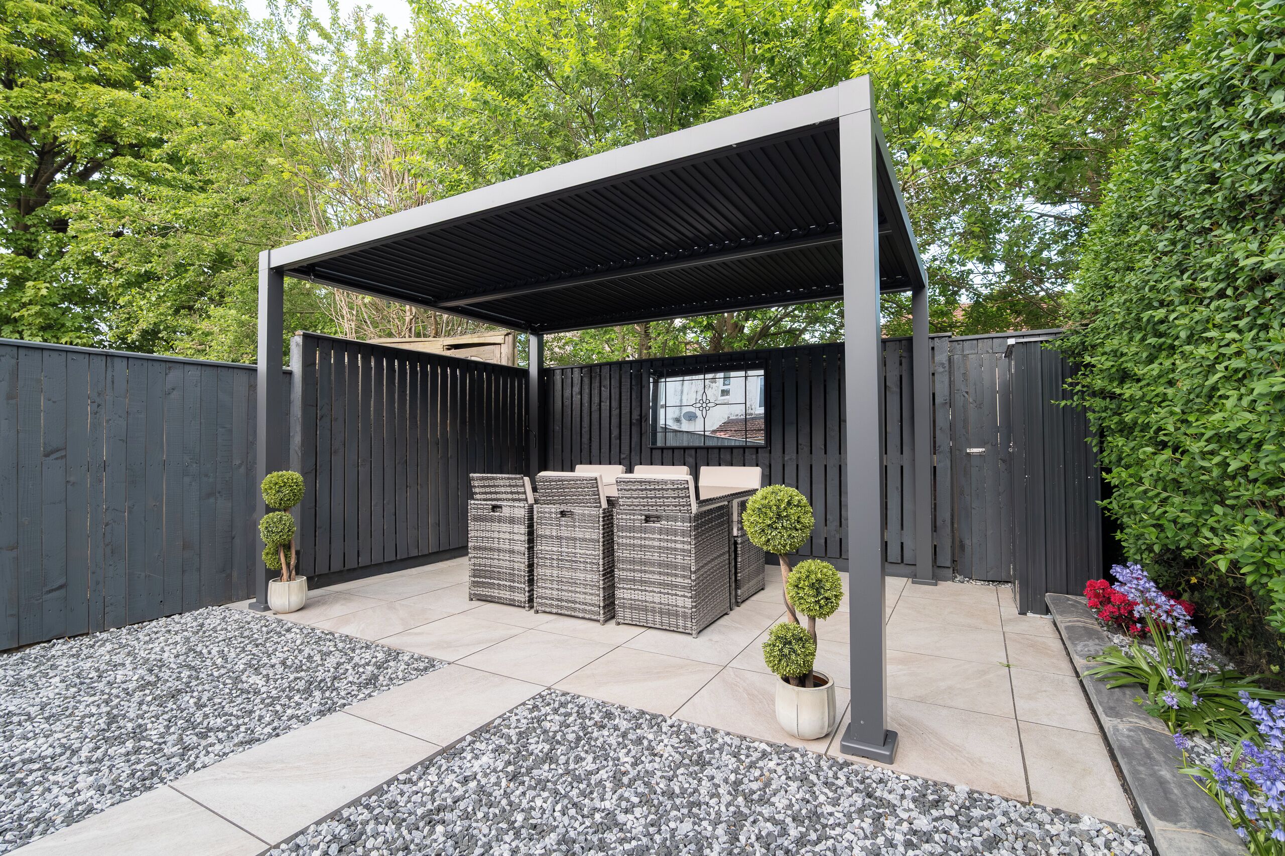 Uma área de varanda coberta com móveis de exterior, uma pérgula e um jardim com plantas e flores.