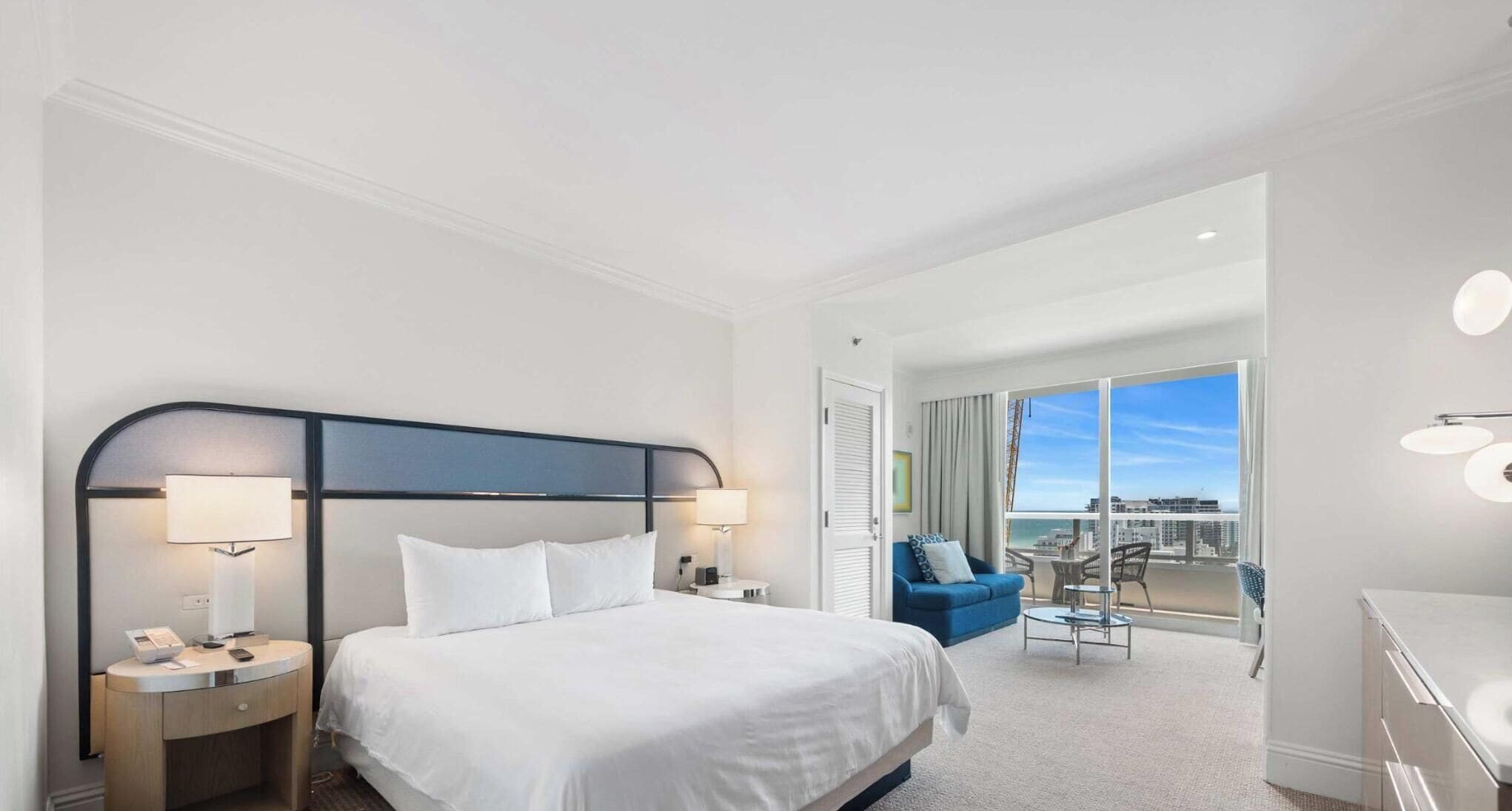 A hotel room with a bed, bedside lamps, a desk, a chair, and a balcony with a view.