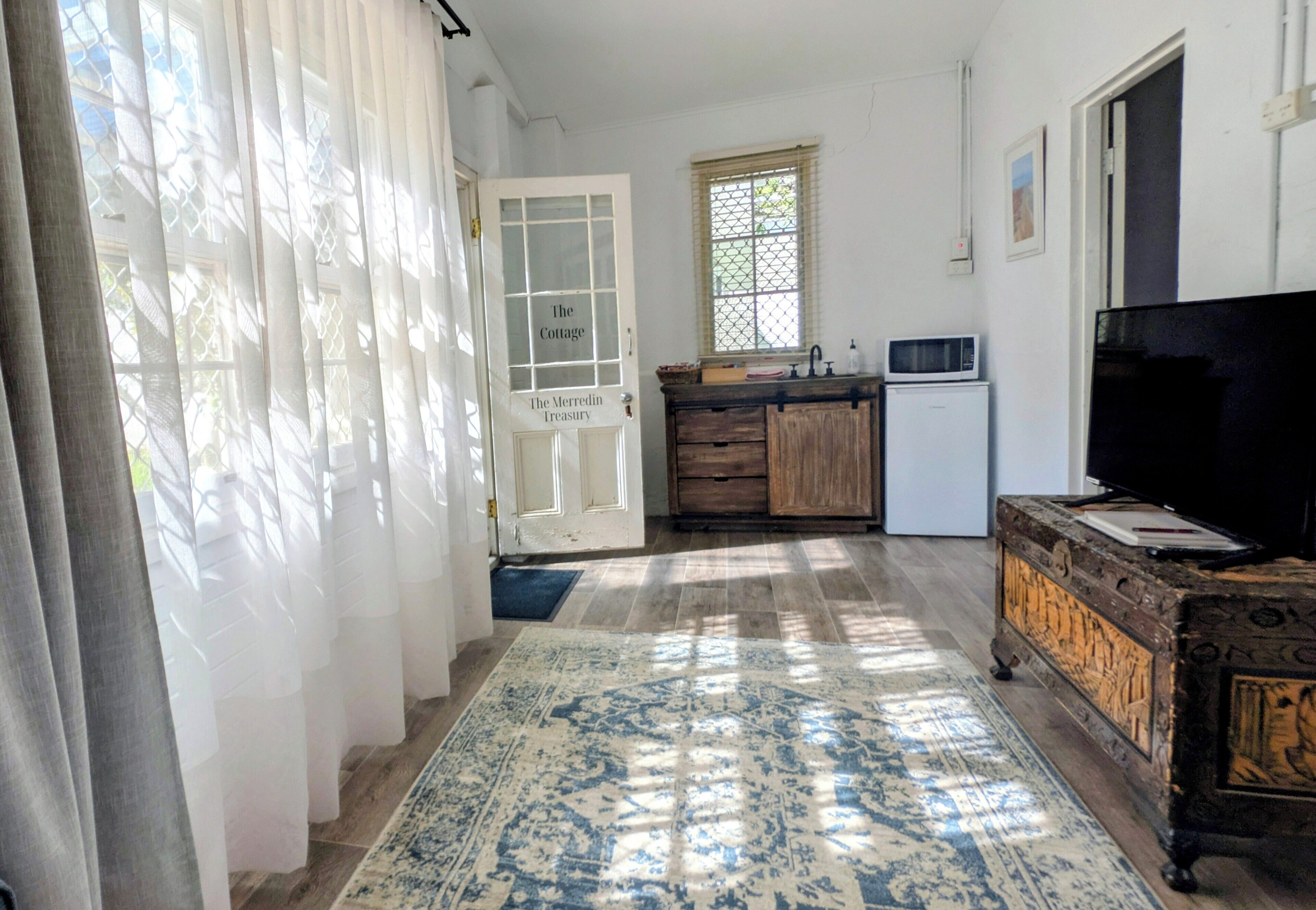 A room with a wooden cabinet, a television, a small refrigerator, a window with a view, and a door labeled 'The Cottage.'