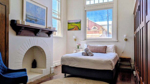 A bedroom with a fireplace, a bed, a chair, and two windows. - The Merredin Treasury (Merredin)