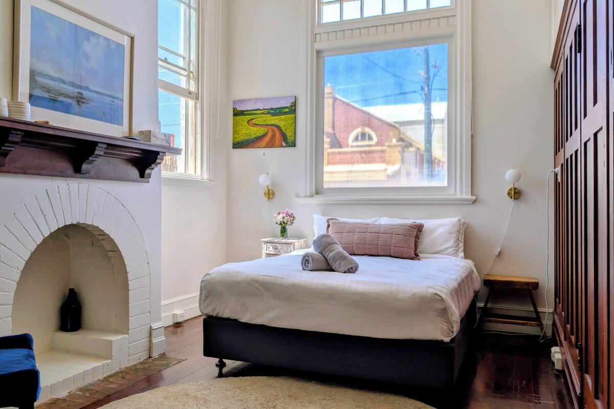 A bedroom with a fireplace, a bed, a chair, and two windows.