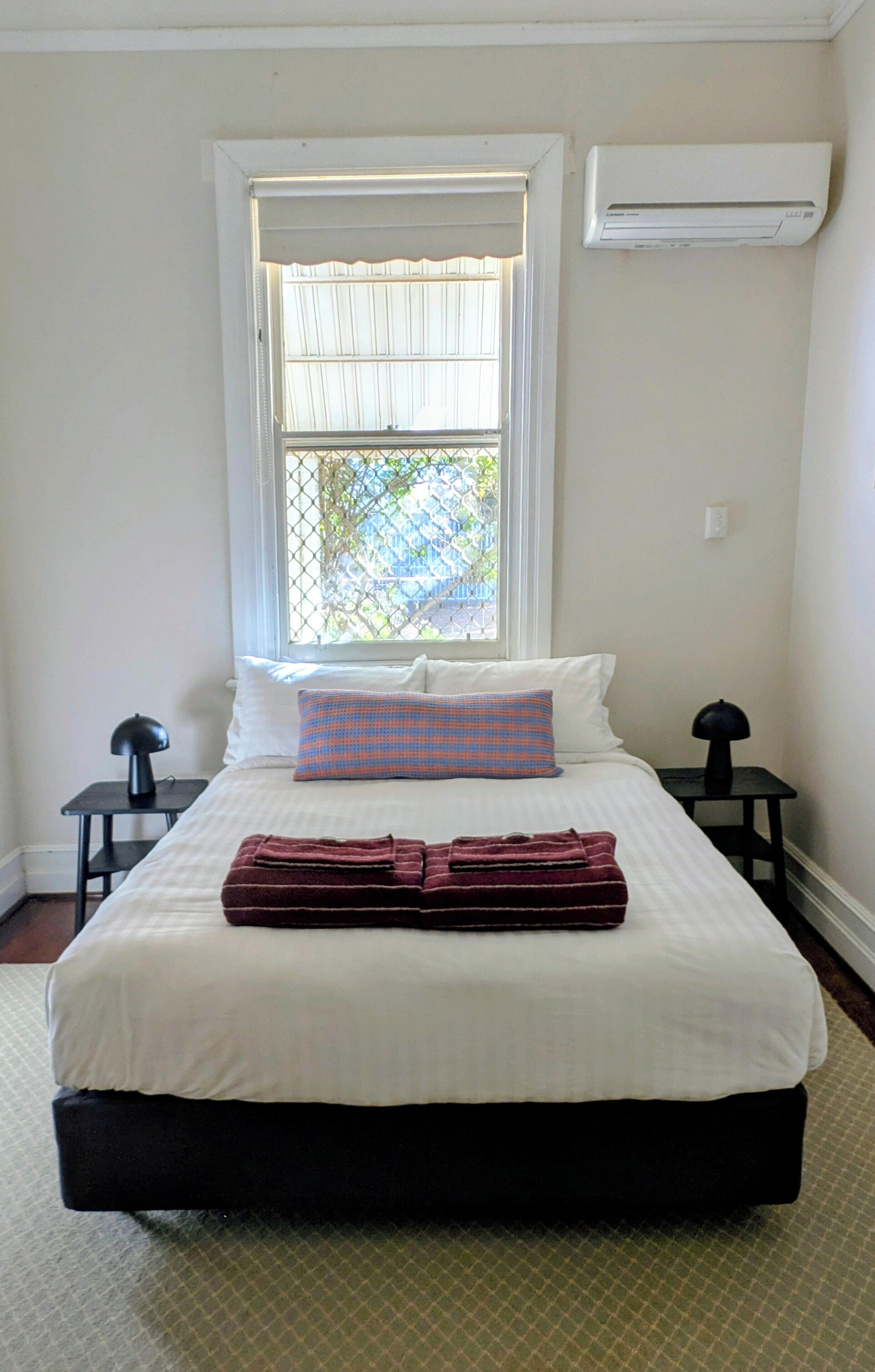 A bedroom with a bed, a window with blinds, an air conditioner, and two nightstands.